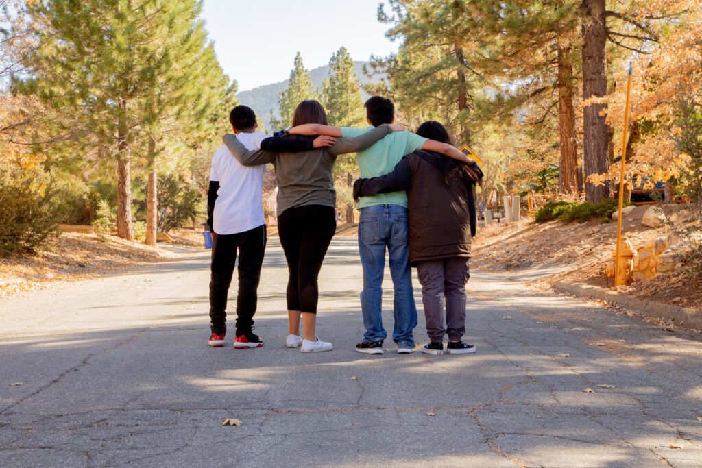 Image of four young people standing in the middle of an empty forest road with their arms around each other